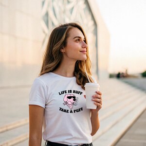 Pu&ograve; includere: T-shirt bianco con grafica rosa di un cane con occhiali da sole e bandana, che fuma una sigaretta. Il testo "LIFE IS RUFF TAKE A PUFF" &egrave; sopra e sotto l'illustrazione del cane. Una persona tiene in mano una tazza di caff&egrave; bianca.