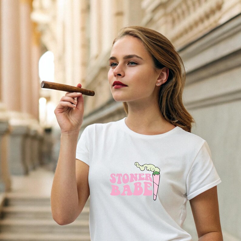 Pu&ograve; includere: T-shirt bianco con la scritta "STONER BABE" in rosa e un'illustrazione di uno spinello acceso. La persona tiene in mano un sigaro. La maglietta &egrave; un capo casual.