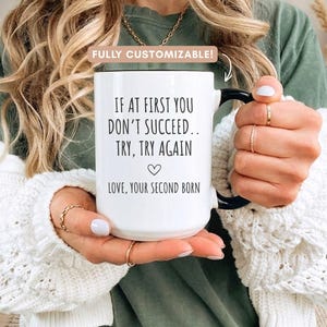 May include: White ceramic mug with a black handle featuring the text "IF AT FIRST YOU DON'T SUCCEED.. TRY, TRY AGAIN" and "LOVE, YOUR SECOND BORN". The mug has a heart symbol and the words "FULLY CUSTOMIZABLE!" above it.