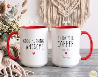 May include: Two white ceramic mugs with bright red interiors and handles. The front mug features the black text GOOD MORNING HANDSOME with a small red heart. The back mug displays ENJOY YOUR COFFEE I LOVE YOU and the name Penelope in black.