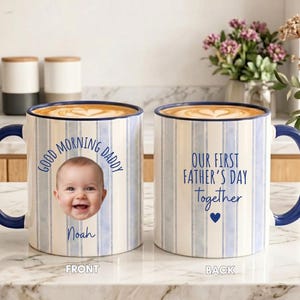 May include: Two ceramic mugs with a blue and white vertical striped pattern and dark blue handles. The front mug features a baby's face, the text "GOOD MORNING DADDY" and "Noah". The back mug displays the text "OUR FIRST FATHER'S DAY together" with a heart.