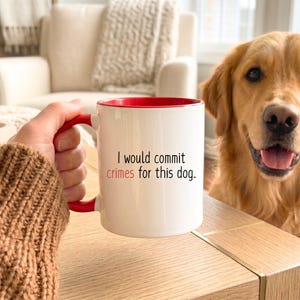 May include: A white ceramic mug with a red interior and handle, featuring the black printed text "I would commit" and the red printed text "crimes" followed by "for this dog." The mug is held by a hand wearing a brown knit sweater sleeve.