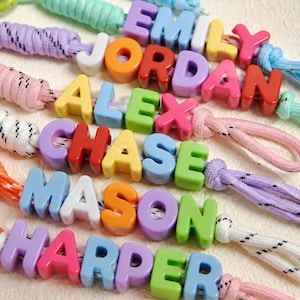 May include: A collection of personalized keychains featuring colorful plastic letter beads spelling out the names Emily, Jordan, Alex, Chase, Mason, and Harper. Each name is attached to a braided nylon cord in various pastel and bright colors.
