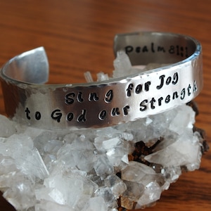 May include: A silver-toned cuff bracelet with the words "Sing for Joy to God our Strength" and "Psalm 81:1" engraved on it. The bracelet has a hammered texture and is displayed on a bed of white crystals.