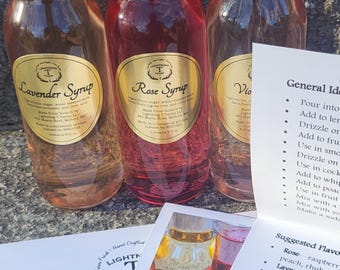 Puede incluir: Tres botellas de vidrio de jarabe con sabor, etiquetadas como "Lavender Syrup", "Rose Syrup" y "Violet Syrup". Cada botella tiene una etiqueta dorada. Una tarjeta de receta y una pequeña imagen de los jarabes están en primer plano.