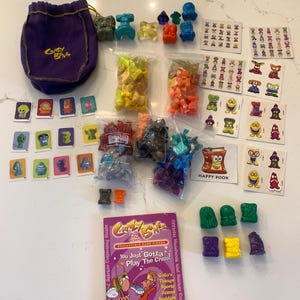 May include: A Crazy Bones collectible game set. The set includes a purple drawstring bag, various colorful game pieces, cards, and a booklet. The game pieces are in various colors and shapes, including green, blue, orange, and purple.