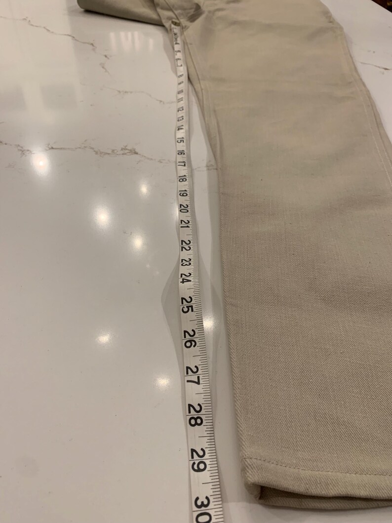 May include: Beige denim jeans with a straight leg, measured with a tape measure. The jeans are approximately 30 inches long. The fabric has a subtle texture, and the color is a light, neutral tone.