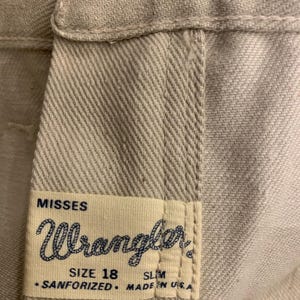 May include: Close-up of light beige Wrangler jeans. The label reads "MISSES Wrangler SIZE 18 SLIM SANFORIZED MADE IN USA." A silver button is visible at the top.