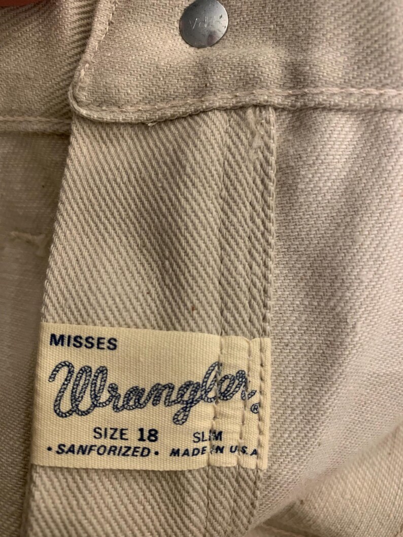 May include: Close-up of light beige Wrangler jeans. The label reads "MISSES Wrangler SIZE 18 SLIM SANFORIZED MADE IN USA." A silver button is visible at the top.