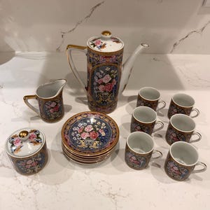 Puede incluir: Juego de té de porcelana vintage con una tetera alta, una jarra de crema, un azucarero, platillos y tazas. El juego tiene un diseño floral con rosas rosas, detalles azules y ribetes dorados.