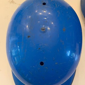 Op de afbeelding: Een bovenaanzicht van een felblauwe honkbalhelm. De helm heeft een afgeronde vorm met een rand en twee kleine gaten aan de bovenkant. Het oppervlak vertoont tekenen van slijtage.