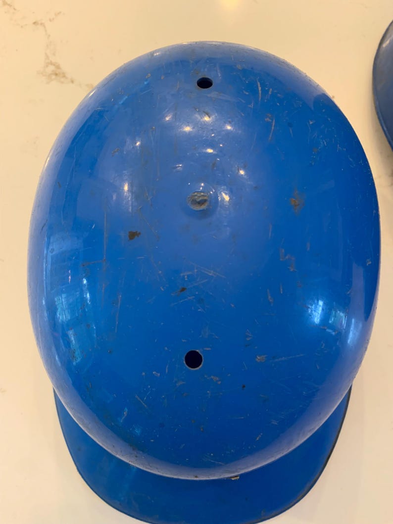 Op de afbeelding: Een bovenaanzicht van een felblauwe honkbalhelm. De helm heeft een afgeronde vorm met een rand en twee kleine gaten aan de bovenkant. Het oppervlak vertoont tekenen van slijtage.