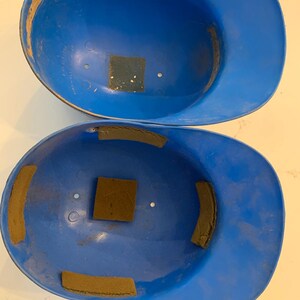 Op de afbeelding: Twee vintage blauwe honkbalhelmen. De helmen zijn felblauw van kleur, met zichtbare slijtage. Elke helm heeft bruine vierkante en rechthoekige vulling aan de binnenkant. De helmen zijn leeg en klaar voor gebruik.