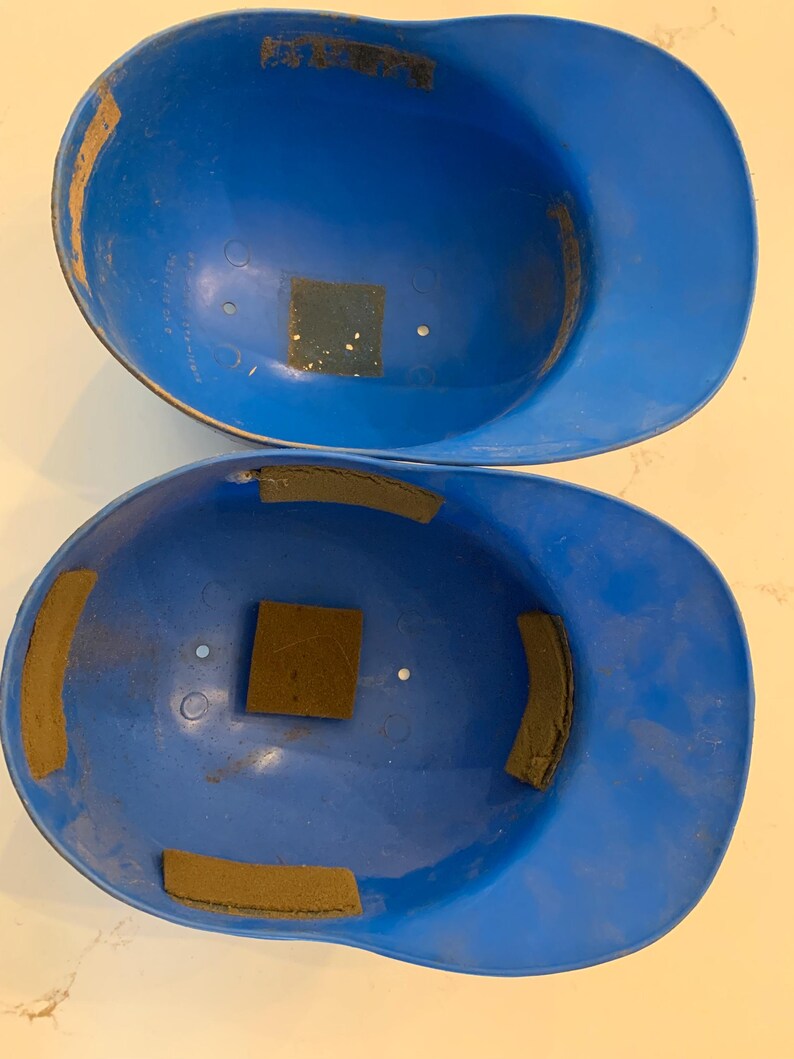Op de afbeelding: Twee vintage blauwe honkbalhelmen. De helmen zijn felblauw van kleur, met zichtbare slijtage. Elke helm heeft bruine vierkante en rechthoekige vulling aan de binnenkant. De helmen zijn leeg en klaar voor gebruik.