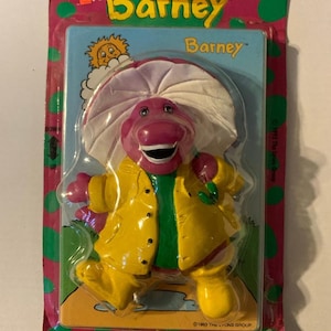May include: A Barney Switchplate toy in its original packaging. The purple dinosaur figure wears a yellow jacket and a white hat. The packaging is pink and green with the Barney logo and the text "Switchplate".