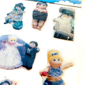 Puede incluir: Una hoja de pegatinas vintage "Fluff Stickers" con varias figuras de muñecas. Las pegatinas representan muñecas con diferentes atuendos, incluyendo una novia y un novio, y otros personajes con vestidos y trajes de baño. La hoja de pegatinas tiene un diseño de arcoíris y nubes.