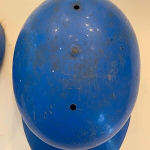 Op de afbeelding: Een blauwe honkbalhelm met een afgeronde bovenkant en een kleine klep. De helm heeft twee kleine gaten aan de bovenkant en vertoont tekenen van slijtage, waaronder krassen en verkleuring. De helm is effen, levendig blauw.