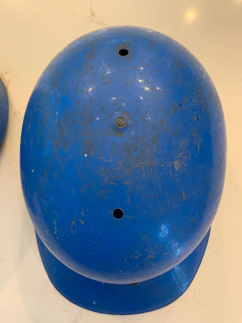 Op de afbeelding: Een blauwe honkbalhelm met een afgeronde bovenkant en een kleine klep. De helm heeft twee kleine gaten aan de bovenkant en vertoont tekenen van slijtage, waaronder krassen en verkleuring. De helm is effen, levendig blauw.