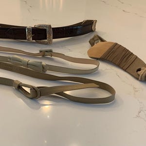 May include: An assortment of belts and a leather knife sheath. The belts include a dark brown embossed leather belt with a silver buckle, and two thin, light-coloured leather belts. The knife sheath is tan and brown.