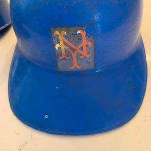 Op de afbeelding: Een vintage blauwe honkbalhelm met het logo van de New York Mets. Het logo is een gestileerde "NY" in oranje en blauw. De helm vertoont tekenen van slijtage.