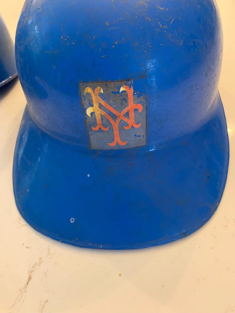 Op de afbeelding: Een vintage blauwe honkbalhelm met het logo van de New York Mets. Het logo is een gestileerde "NY" in oranje en blauw. De helm vertoont tekenen van slijtage.