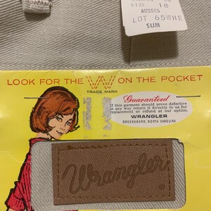 May include: Close-up of light gray denim jeans with a tag. The tag indicates the jeans are 100% cotton, size 18 misses, and slim fit. A vintage Wrangler label is visible, along with the text "Look for the W on the pocket."