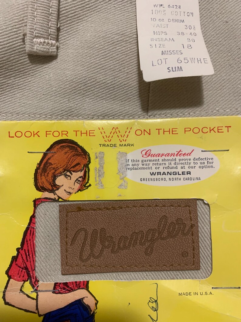 May include: Close-up of light gray denim jeans with a tag. The tag indicates the jeans are 100% cotton, size 18 misses, and slim fit. A vintage Wrangler label is visible, along with the text "Look for the W on the pocket."