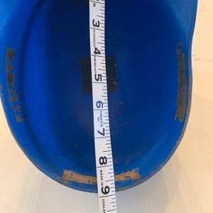 Op de afbeelding: Een felblauwe honkbalhelm met een meetlint dat een lengte van ongeveer 23 cm aangeeft. De helm heeft wat slijtage aan de randen. Het meetlint is wit met zwarte markeringen.