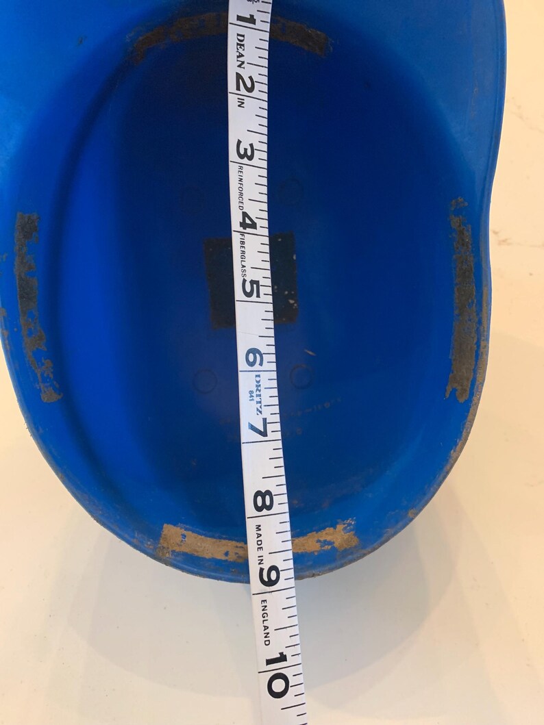 Op de afbeelding: Een felblauwe honkbalhelm met een meetlint dat een lengte van ongeveer 23 cm aangeeft. De helm heeft wat slijtage aan de randen. Het meetlint is wit met zwarte markeringen.