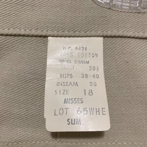 May include: Close-up of a tag attached to beige denim pants. The tag reads "100% Cotton, 10 oz Denim, Waist 30 1/2, Inseam 30, Size 18, Misses, Lot 65WHE, Slim". The pants are hanging on a clear plastic hanger.