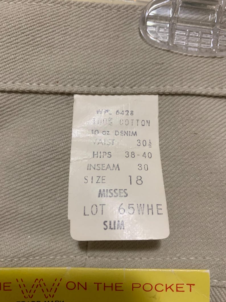 May include: Close-up of a tag attached to beige denim pants. The tag reads "100% Cotton, 10 oz Denim, Waist 30 1/2, Inseam 30, Size 18, Misses, Lot 65WHE, Slim". The pants are hanging on a clear plastic hanger.