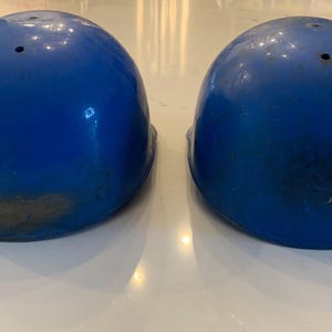 Op de afbeelding: Twee blauwe helmen met een glanzende afwerking. Elke helm heeft een enkel ventilatiegat aan de bovenkant en vertoont tekenen van slijtage. De helmen zijn waarschijnlijk voor bescherming.