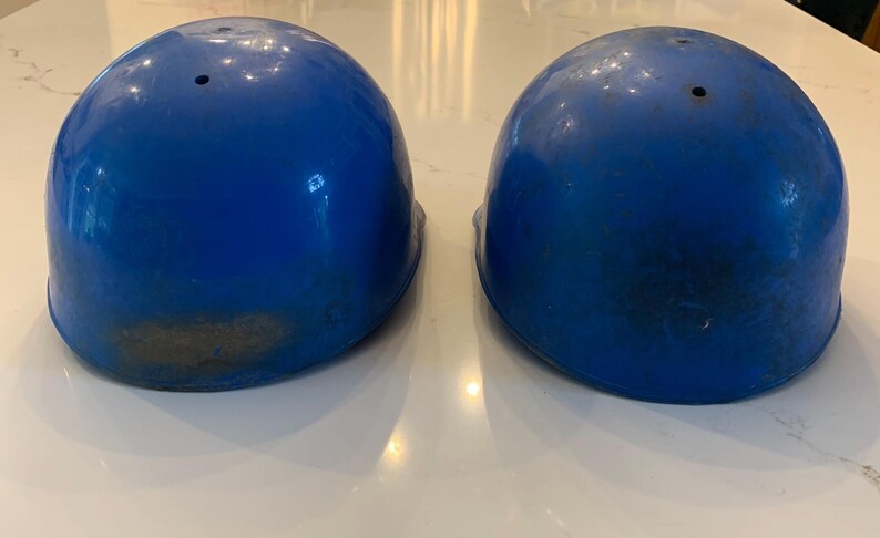 Op de afbeelding: Twee blauwe helmen met een glanzende afwerking. Elke helm heeft een enkel ventilatiegat aan de bovenkant en vertoont tekenen van slijtage. De helmen zijn waarschijnlijk voor bescherming.