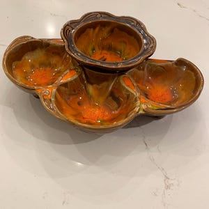 Puede incluir: Una fuente de cerámica con cuatro secciones. La fuente tiene un exterior marrón con interior naranja y amarillo. El cuenco central es más pequeño y tiene un borde azul oscuro. Los otros tres cuencos son más grandes y tienen un diseño ondulado.