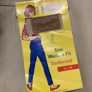 May include: Tan-colored pants with a yellow tag featuring a vintage illustration of a woman in blue jeans and a red shirt. The tag reads "Wrangler" and "Trim Western Fit Sanforized SLIM."
