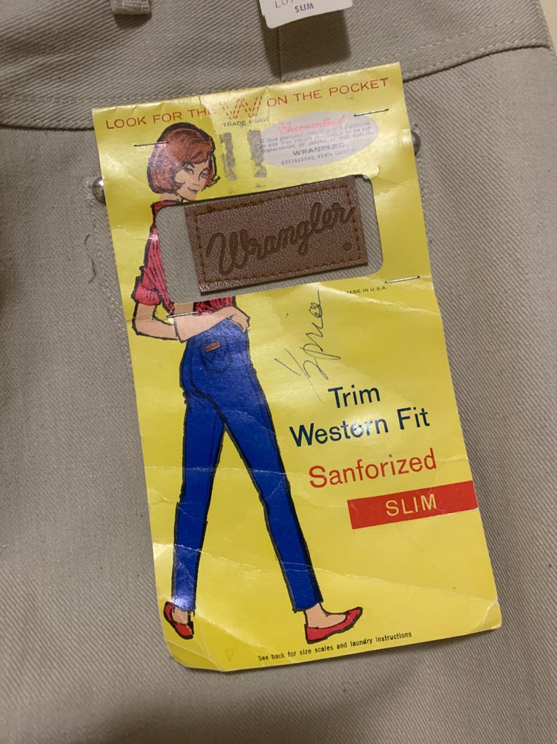 May include: Tan-colored pants with a yellow tag featuring a vintage illustration of a woman in blue jeans and a red shirt. The tag reads "Wrangler" and "Trim Western Fit Sanforized SLIM."