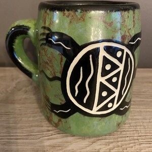 May include: A ceramic mug with a mottled green and brown glaze featuring a prominent black and white graphic of a turtle on the side. The turtle shell is decorated with geometric patterns including triangles and dots, and the handle is black.