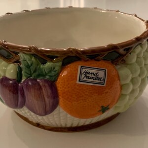 May include: A ceramic fruit bowl with a cream interior and a basket weave exterior. The bowl is decorated with grapes, plums, and an orange. The rim is brown and vine-like. A sticker on the orange reads "Hand Painted."
