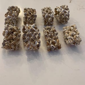 May include: A collection of nine napkin rings, each crafted from small, natural seashells. The shells are primarily beige and brown, creating a textured, organic appearance. These decorative rings are arranged on a light-colored surface.
