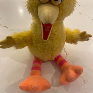 May include: A plush toy of a yellow bird character with a fuzzy body, large eyes with blue and pink eyelids, and an open beak. The toy has long legs with horizontal pink and orange stripes and large, flat orange feet extending outward.