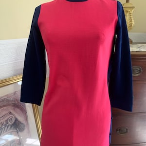 May include: A vintage-style tunic top in a vibrant red with navy blue sleeves and trim. The top features a ribbed texture on the front and three-quarter sleeves. Buttons adorn the shoulder and side seams, adding a classic touch.