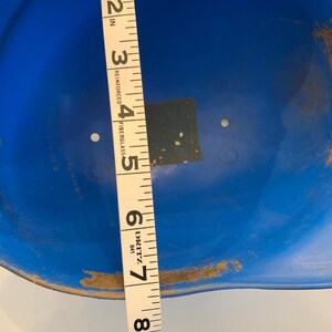 Op de afbeelding: Een blauwe helm met een meetlint dat een diameter van ongeveer 20 cm aangeeft. De helm is gemaakt van glasvezel en heeft wat slijtage aan de randen.