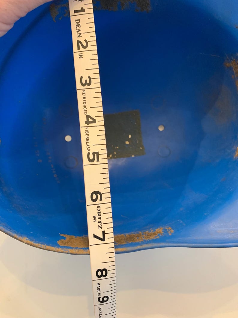 Op de afbeelding: Een blauwe helm met een meetlint dat een diameter van ongeveer 20 cm aangeeft. De helm is gemaakt van glasvezel en heeft wat slijtage aan de randen.
