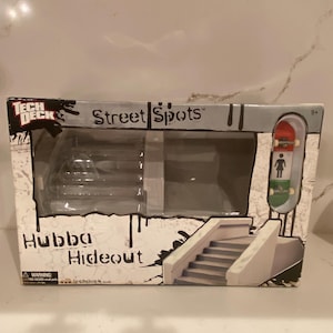 May include: A Tech Deck Street Spots Hubba Hideout fingerboard playset in its original packaging. The box features a white and black graphic design, a clear plastic window, and includes a miniature skateboard with a red, white, and green deck.
