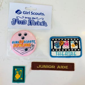May include: A collection of four Girl Scouts iron-on patches including a round pink bowling patch, a rectangular theatre patch with film strip design, a small teal patch with a yellow planet, and a brown rectangular patch labeled JUNIOR AIDE.