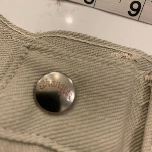 May include: Close-up of a beige denim garment with a silver button featuring the brand name "Wrangler" in red lettering. A measuring tape shows the 6-9 inch range. The garment's stitching is visible.