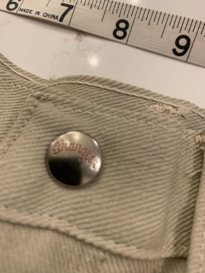May include: Close-up of a beige denim garment with a silver button featuring the brand name "Wrangler" in red lettering. A measuring tape shows the 6-9 inch range. The garment's stitching is visible.