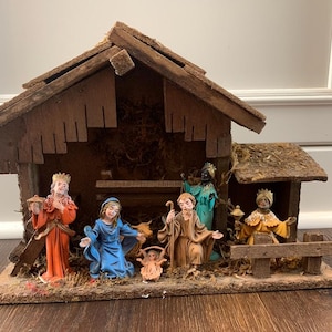 Op de afbeelding: Een rustieke houten kerststal met beeldjes. De scène omvat een stal, Maria, Jozef, het kindje Jezus en de drie wijzen. De beeldjes zijn geschilderd in levendige kleuren, met gouden accenten.