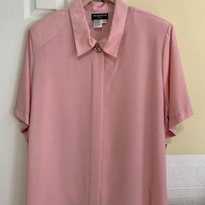 Könnte beinhalten: Eine rosa Bluse mit kurzen Ärmeln und Knopfleiste. Die Bluse hat einen goldenen Knopf vorne und das Label "Marathon" am Kragen.