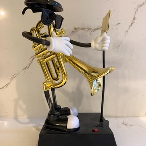 May include: A gold-colored trumpet-shaped figurine with black sunglasses, a black hat, and white gloves. The figurine is on a black base with a red switch and white feet. It holds a gold-colored music sheet.